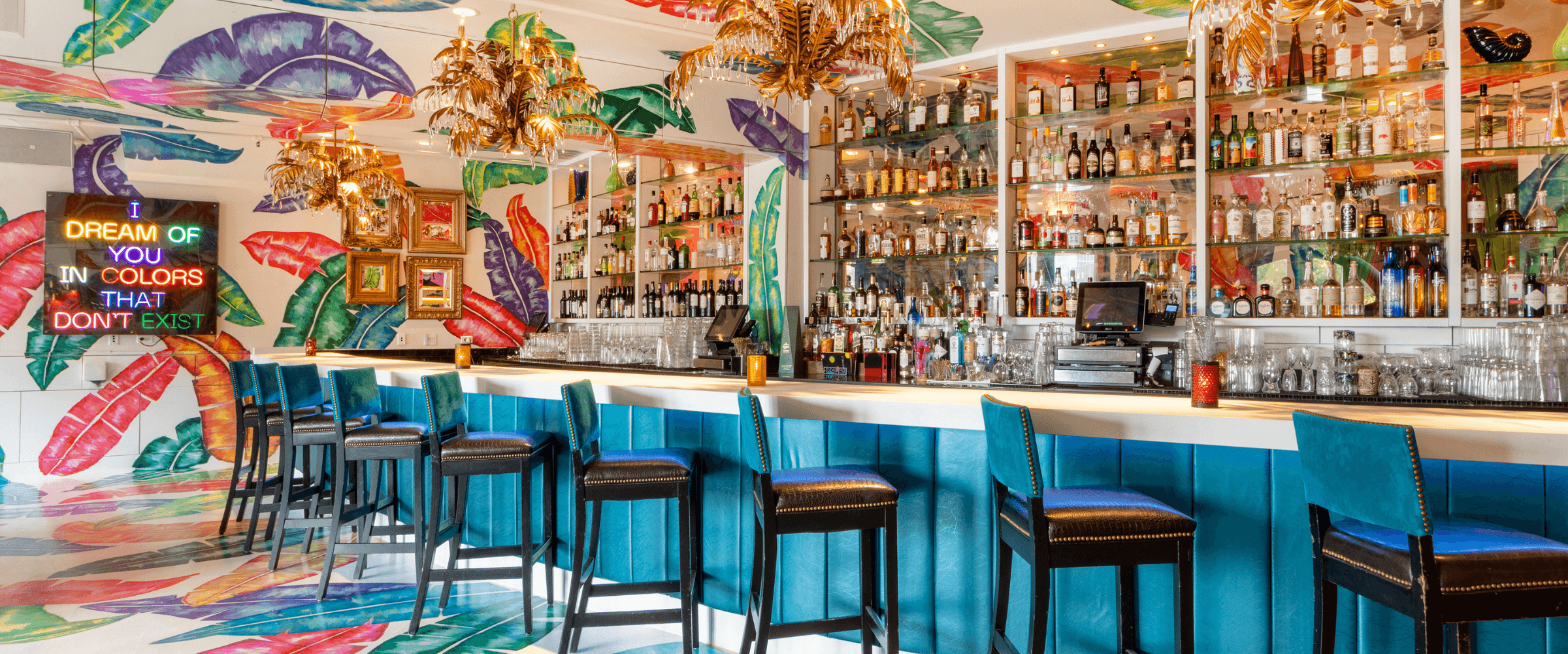 Colorful interior of the Rumba Lounge at Carnivale Chicago, featuring tropical leaf murals, turquoise bar seating, gold chandeliers, a fully stocked bar, and a vibrant neon sign. - Desktop